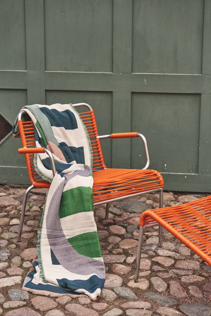 Siirto pläd är formgiven av Maija Isola för Marimekko och har ett distinkt mönster. Här hängd över orange spaghetti-stol.