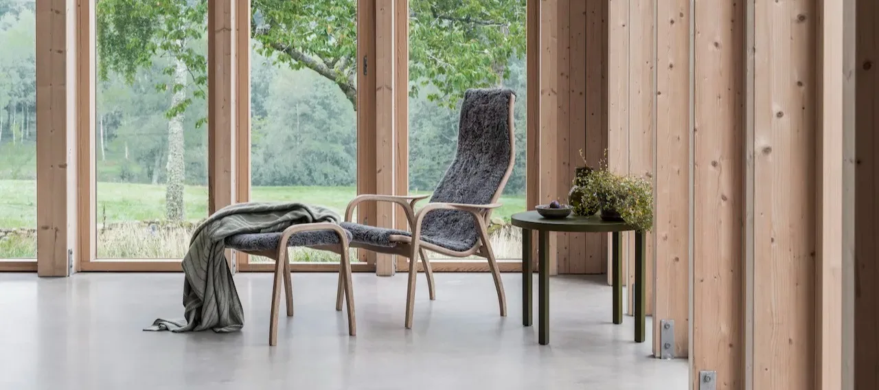 Lamino fåtölj och fotpall i grått fårskinn, formgiven av Yngve Ekström. Här placerad i ljust vardagsrum med stora fönsterpartier.