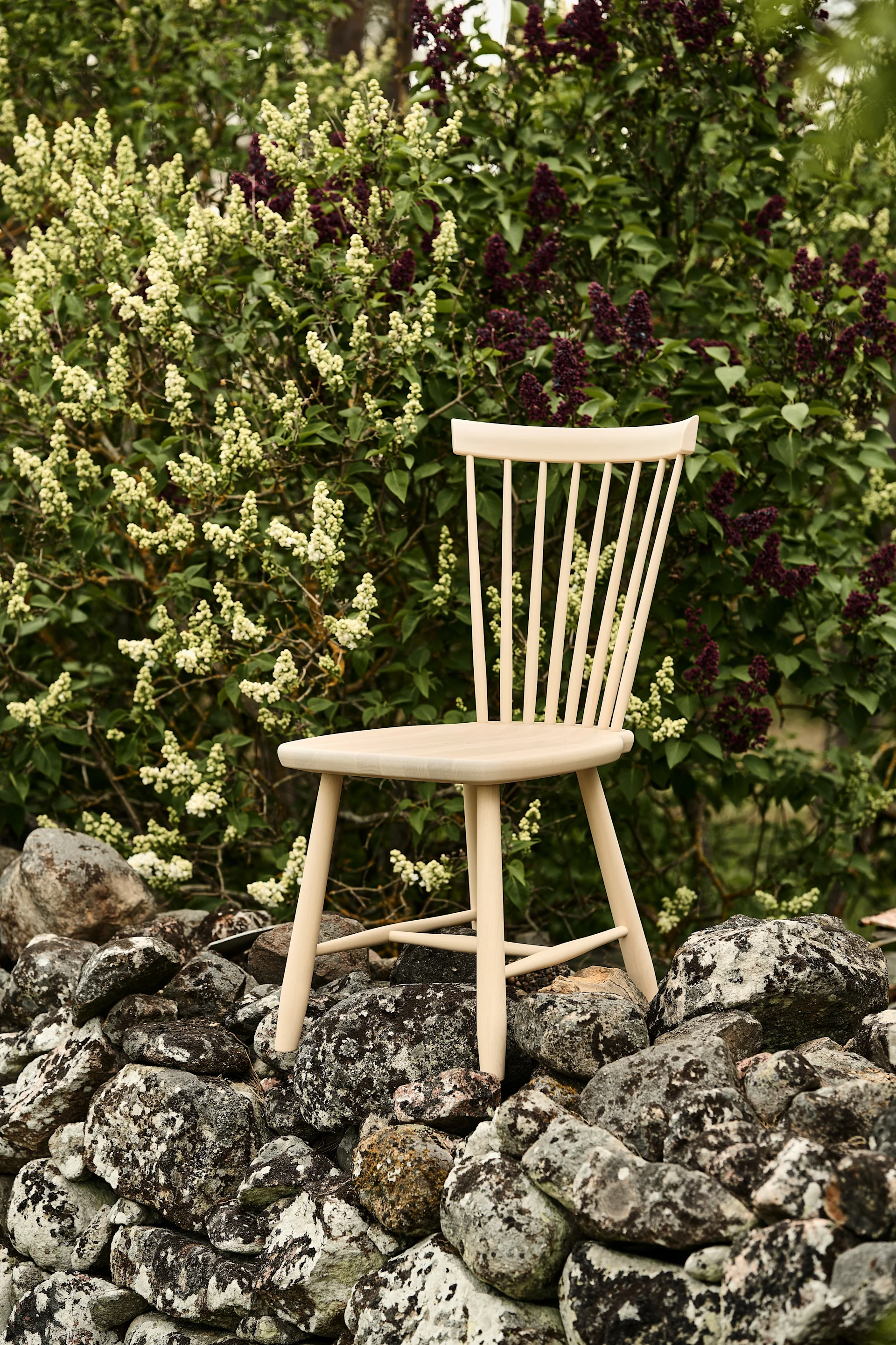 Bild som visar Lilla Åland stol i björk, placerad på stenmur med blommande syrénhäck bakom.