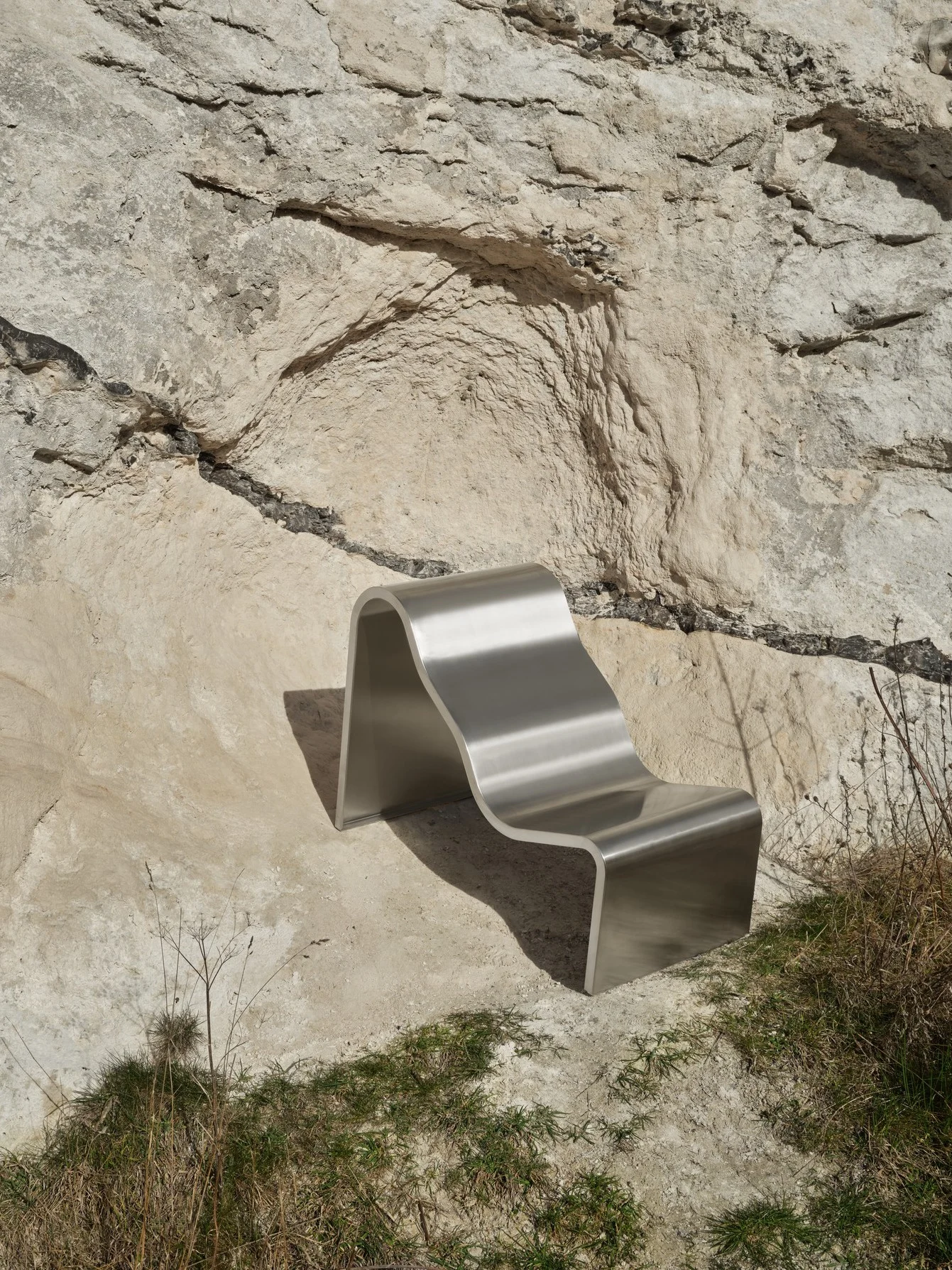 En modern, vågformad silverbänk står mot en ljus, stenig klippvägg med torrt gräs i förgrunden.