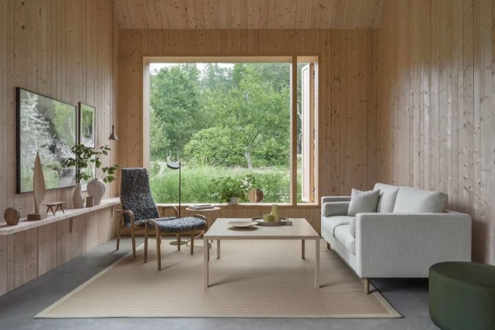 Lamino fåtölj i grått fårskinn och oljad ek med tillhörande fotpall, formgivna av Yngve Ekström för Swedese. Här placerade i vardagsrum med furuklädda väggar och stort fönsterparti.