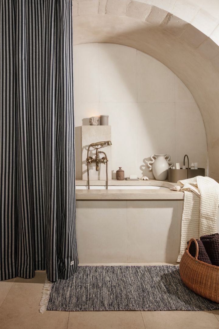 Bild som visar Chambray duschdraperi från Ferm Living, ett randigt duschdraperi i grått och blått placerat i ett litet badrum med badkar. På golvet ligger en grå matta och en stor korg i rotting skymtar i hörnet.