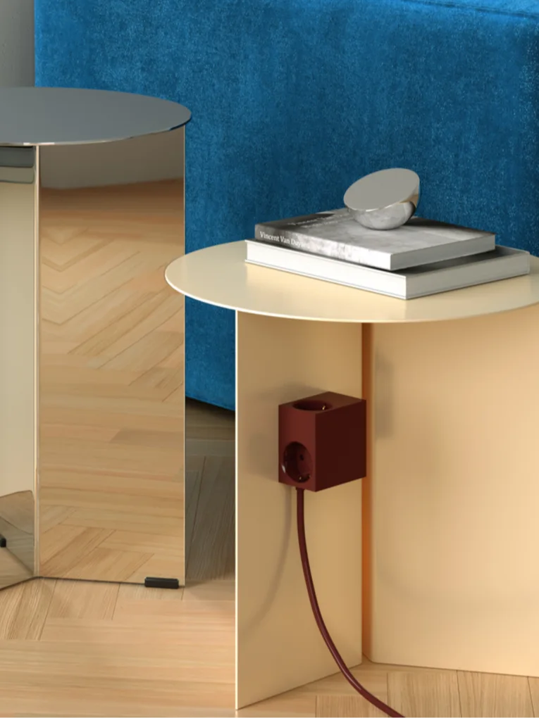 Två moderna sidobord. Ett spegelblankt och ett beige, med böcker, en silverdekoration och en röd strömkub. Blått tyg bakom.