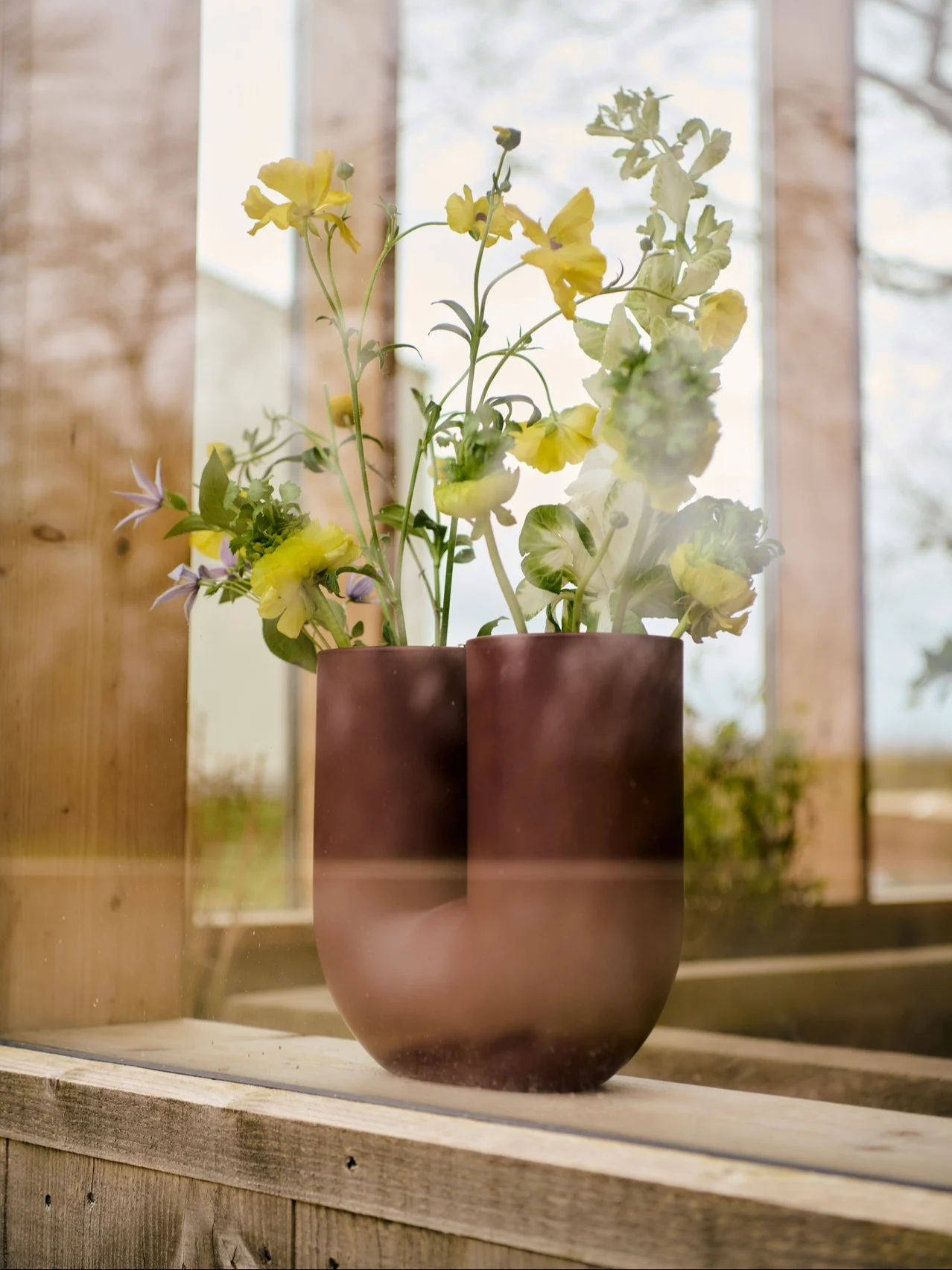 Mörkbrun U-formad vas med gula, ljusgröna och lila blommor på en fönsterbräda i trä.