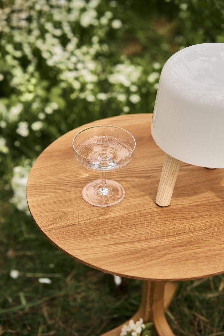 Bild som visar Ripple champagneglas från Ferm Living, placerad på ett Lamino-bord i ek från Swedese på en blomstrande äng.
