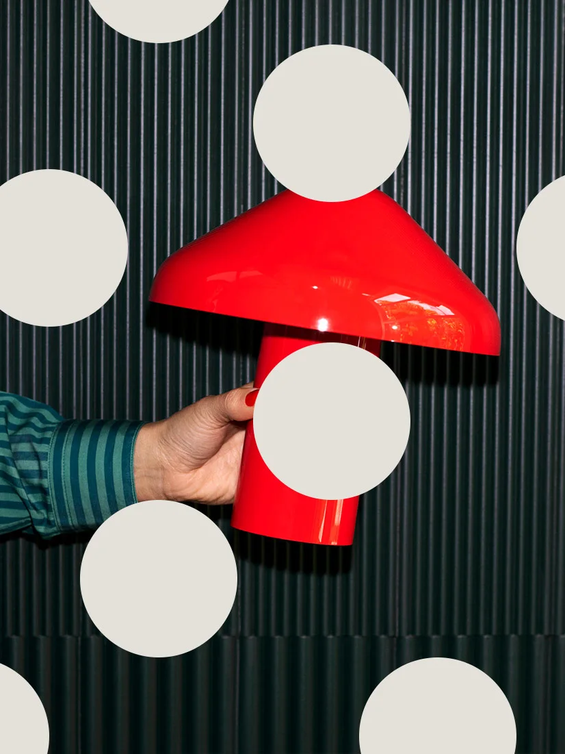 Hand håller en knallröd, svampformad bordslampa mot en mörk räfflad bakgrund.