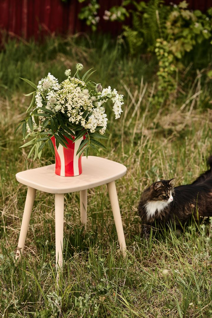 Bild som visar Sobremesa Stripe vas från HAY, en röd- & vitrandig vas placerad på Palle pall från Stolab. I vasen står en bukett med sommarblommor.