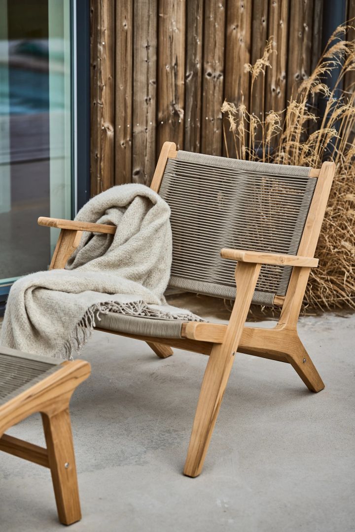 Bild som visar Sandvik loungefåtölj i teak och rep av polypropen. Stolen står placerad på stenlagt uteplats framför ett mörkt trähus, på loungefåtöljens armstöd hänger en beige pläd.