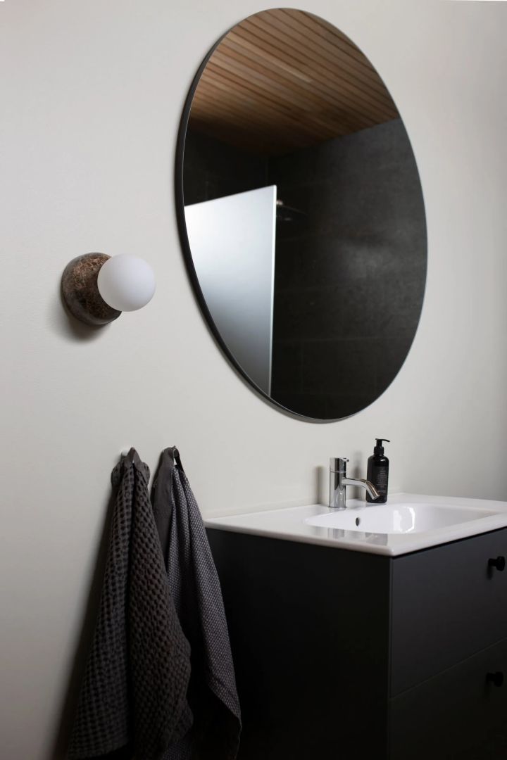 Bild som visar lyxigt badrum med ljusgråa väggar och svart kommod. På väggen sitter den runda lampan Torrano med sockel i brun marmor och klotformad lampa från Globen Lighting.