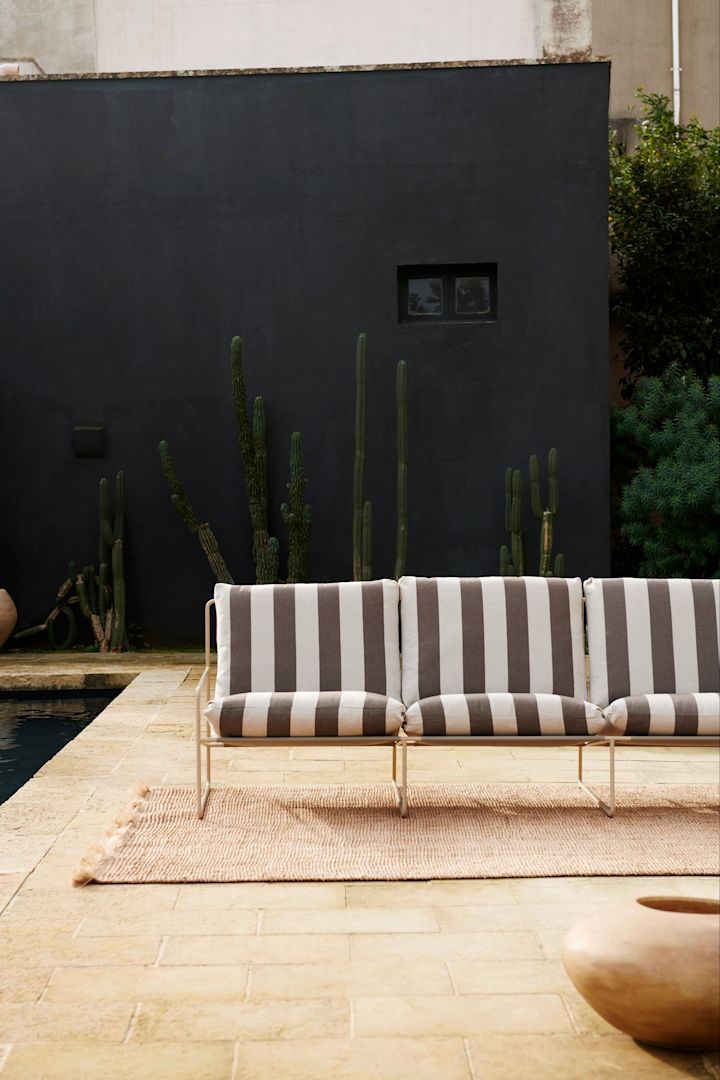 Bild som visar Desert loungesoffa från Ferm Living i ett grå och vit-randigt tyg. Soffan står på en ljus matta framför ett svart hus på stenlagd uteplats.
