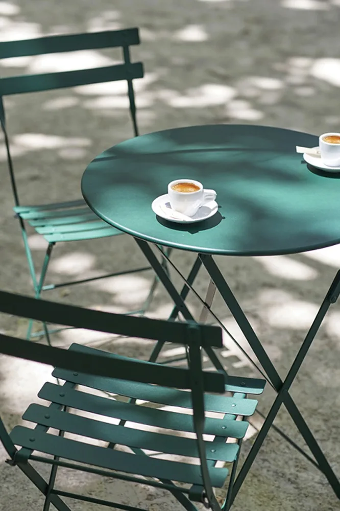 Miljöbild föreställandes bistro cafébord och stolar i färgen cedar green. ståendes på en grusplätt och har två kaffekoppar på sig. 