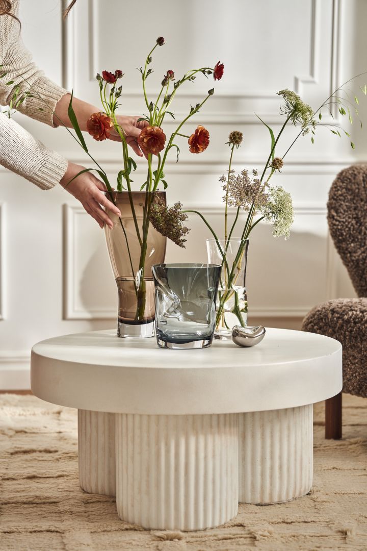 Squeeze vaser i olika storlekar och i färgerna brun, blå och klarglas. Formgivna av Lena Bergström för Orrefors. Här placerade på vitt, runt stenbord.