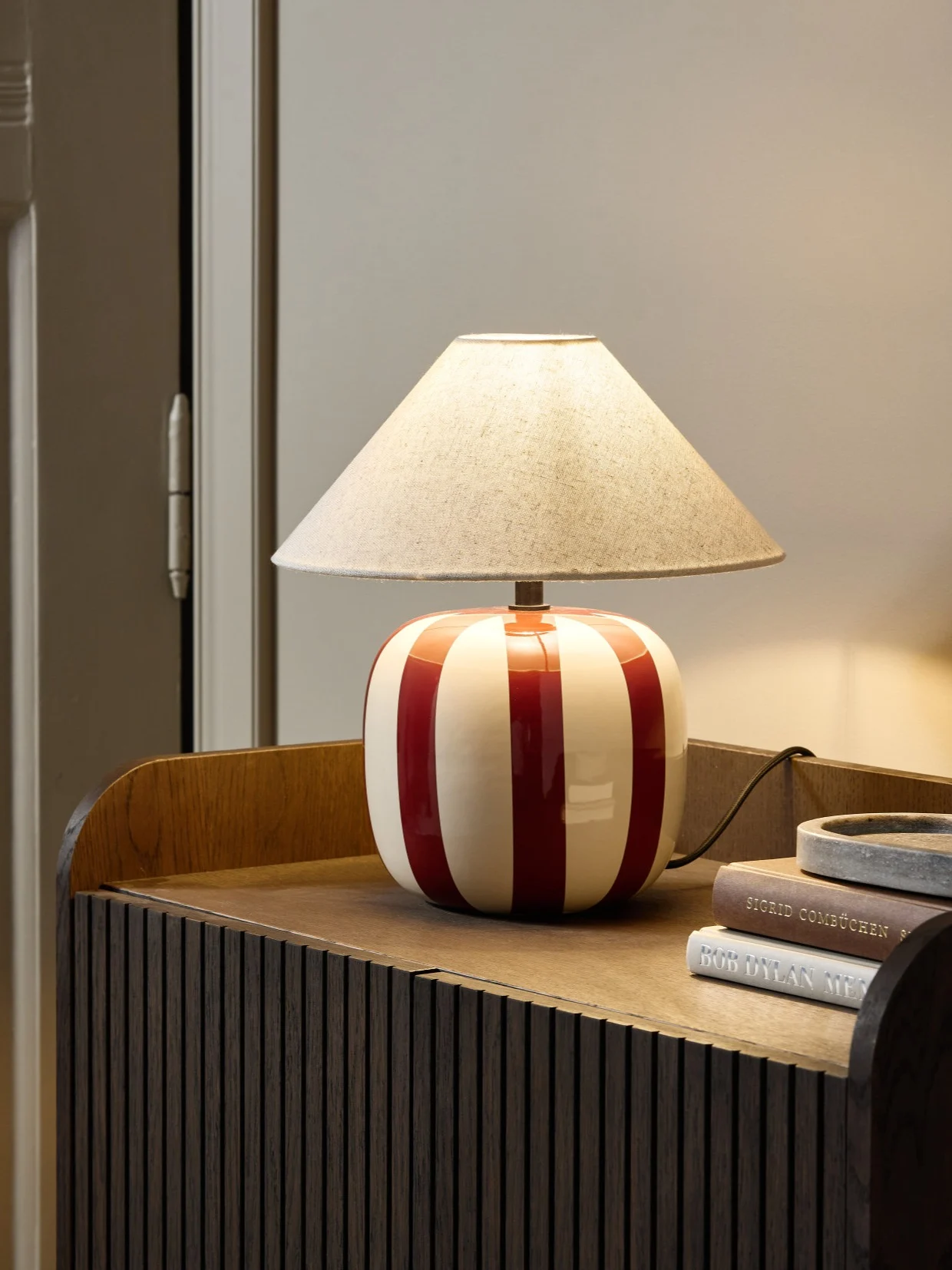 En bordslampa med röd- och vitrandig keramikfot och beige textilskärm står på ett mörkt träskåp.