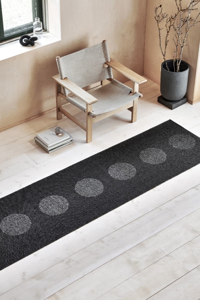 Välj rätt matta som exempelvis en vävd plastmatta, bild som visar Vera plastmatta i svart metallic från Pappelina, placerad på ljust trägolv med en ljus stol och svart golvkruka bredvid.