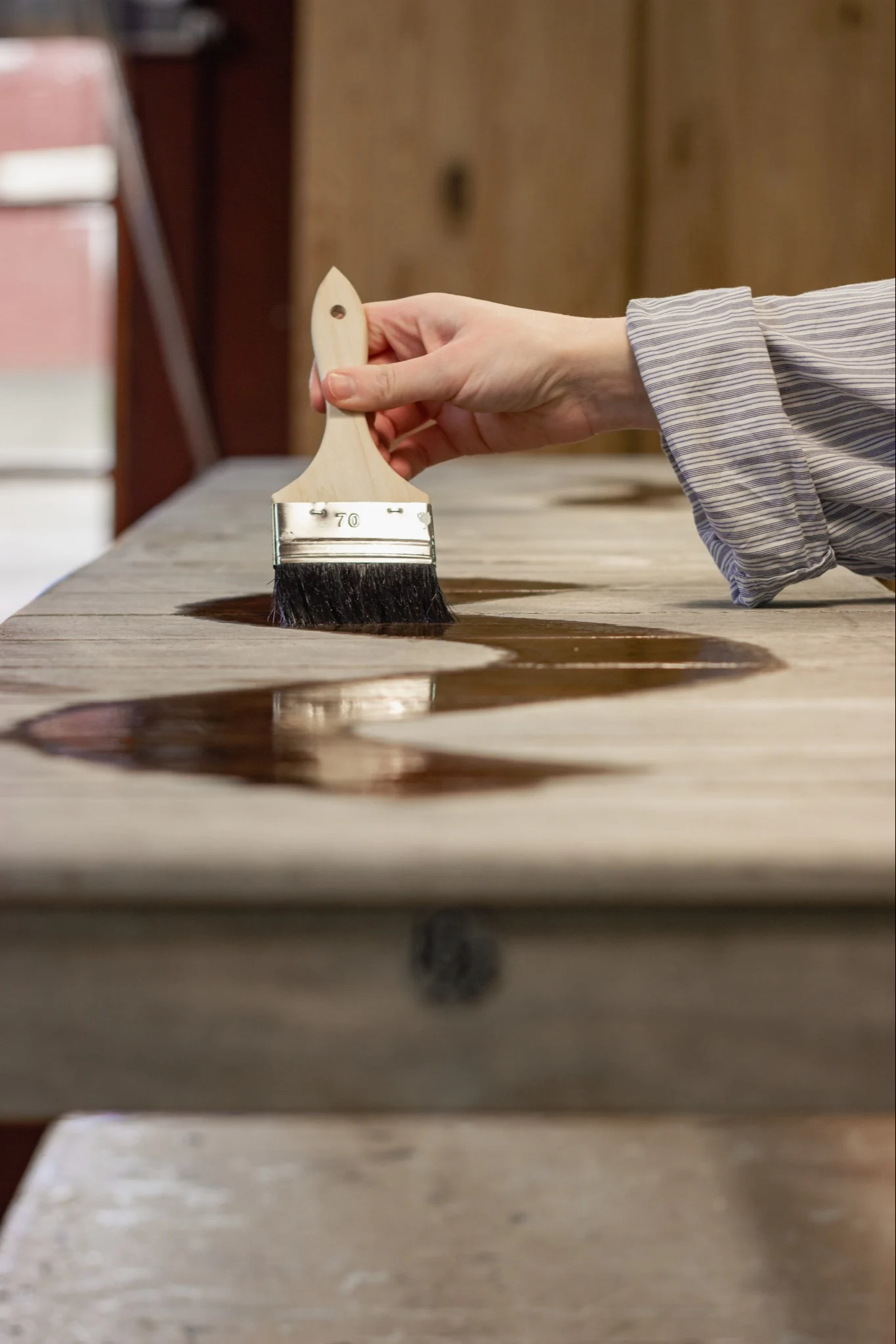 Hand applicerar mörk bets eller lack med pensel på en ljus träyta, vilket skapar ett vågigt mönster.