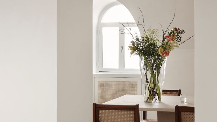 Bild som visar Reed vas 50 cm i klarglas från Orrefors, formgiven av Monica Förster, här placerad på ett matbord och fylld med en spretig bukett där färgglada blommor kombineras med kvistar och strån.