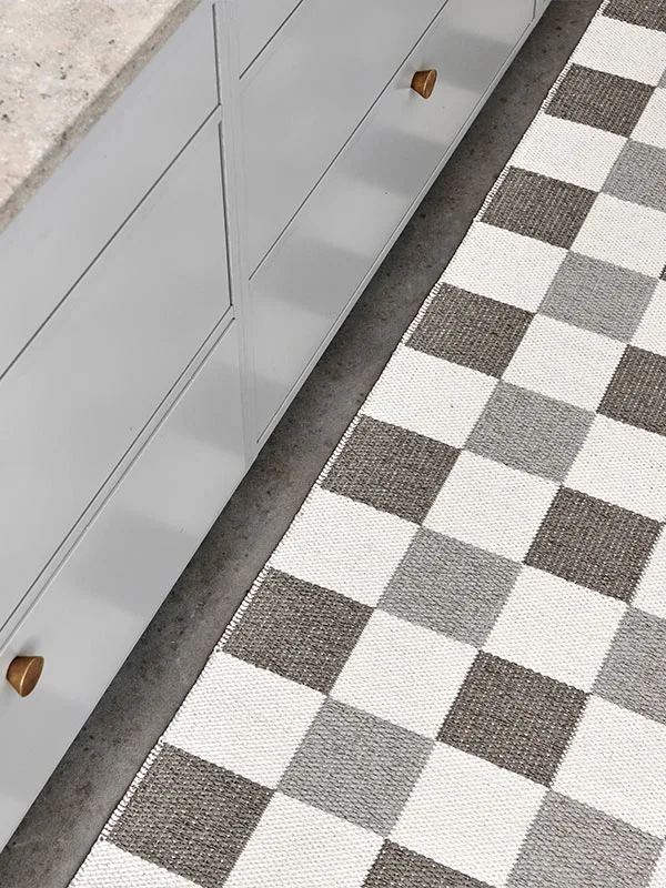 Rutigt matta i grått och vitt bredvid ljusgrå köksskåp med mässingsknoppar och en bänkskiva.