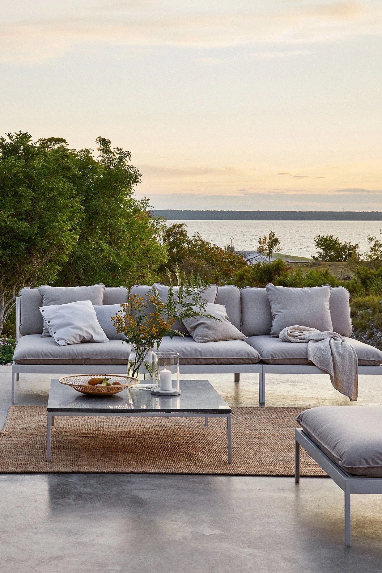Bild som visar Bönan modulsoffa från Skargaarden. Denna modell är i färgen light grey med vitt underrede. Här står loungesoffan placerad på stor uteplats med stengolv, med trädtoppar och havsutsikt i bakgrunden.