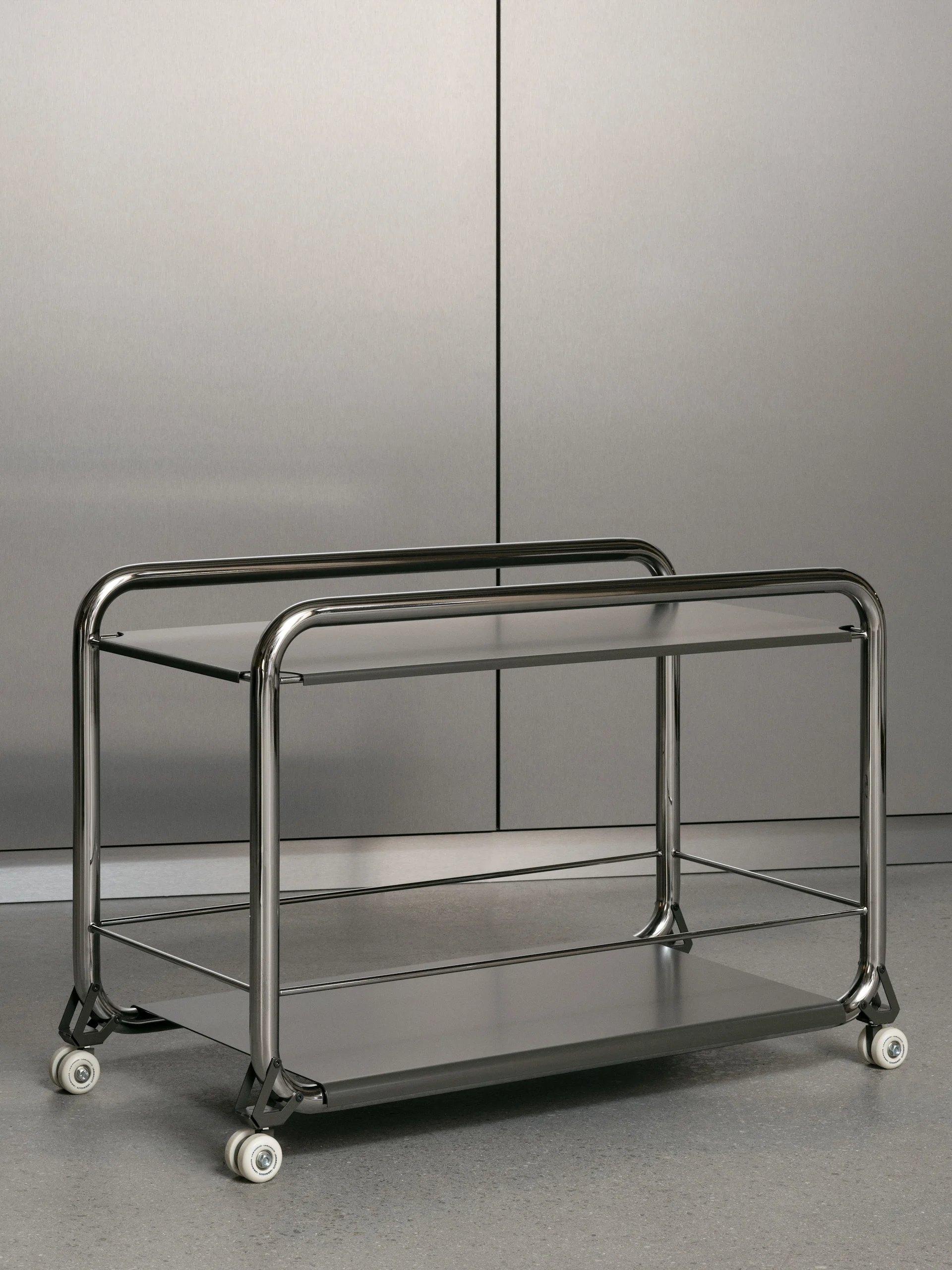 En rullvagn i rostfritt stål med två hyllplan, rundad ram och vita hjul, på ett grått golv framför en vägg i borstad metall.