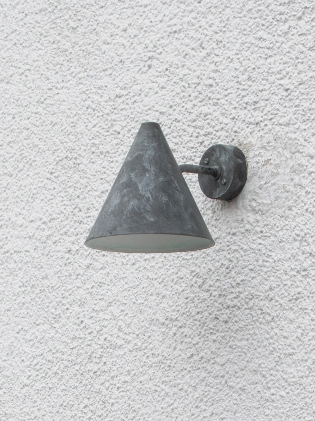 En väderbiten mörkgrå konisk utomhusvägglampa monterad på en strukturerad vit vägg.