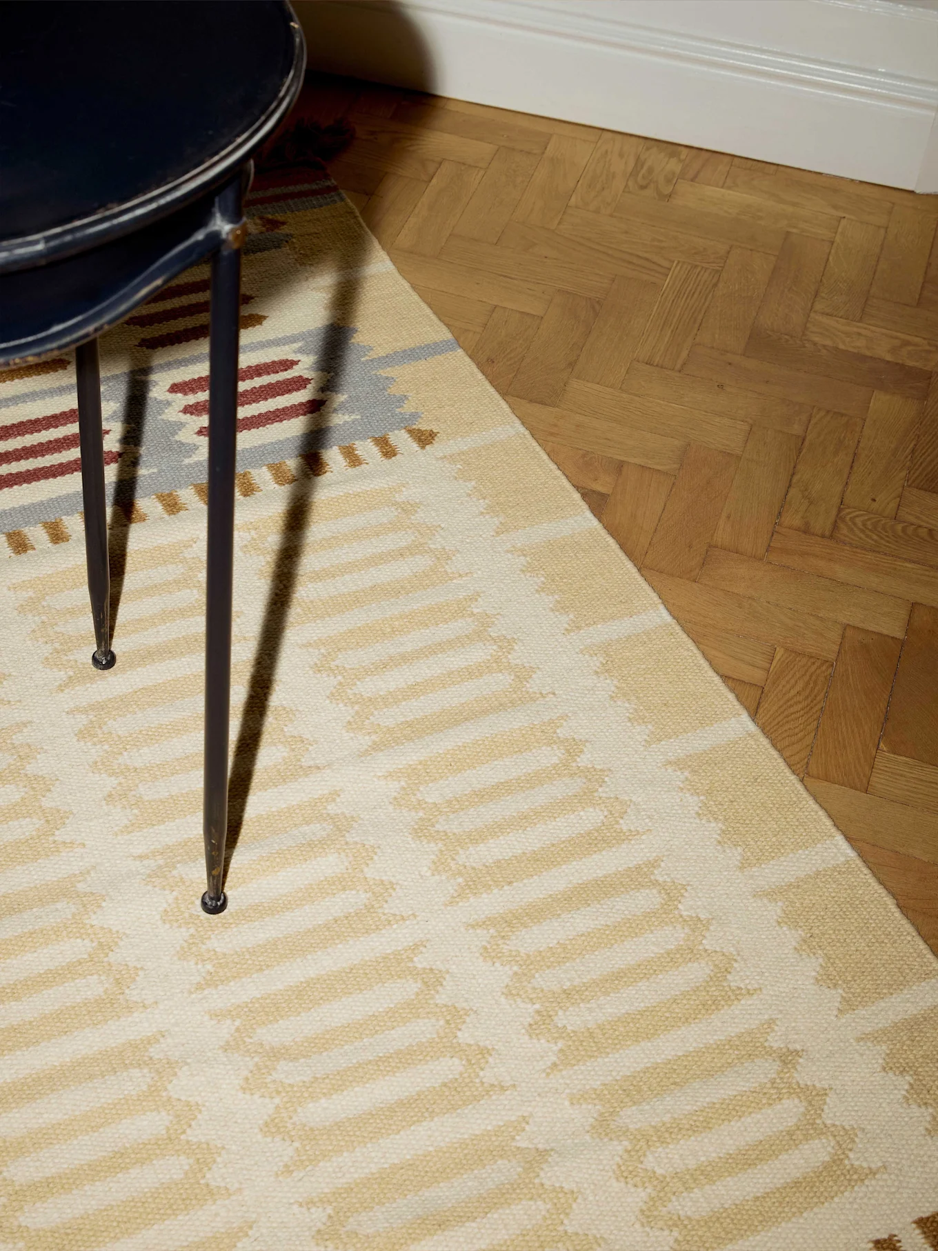 Närbild på en beige matta med geometriska mönster i blått, rött och guld, bredvid ett fiskbensparkettgolv.