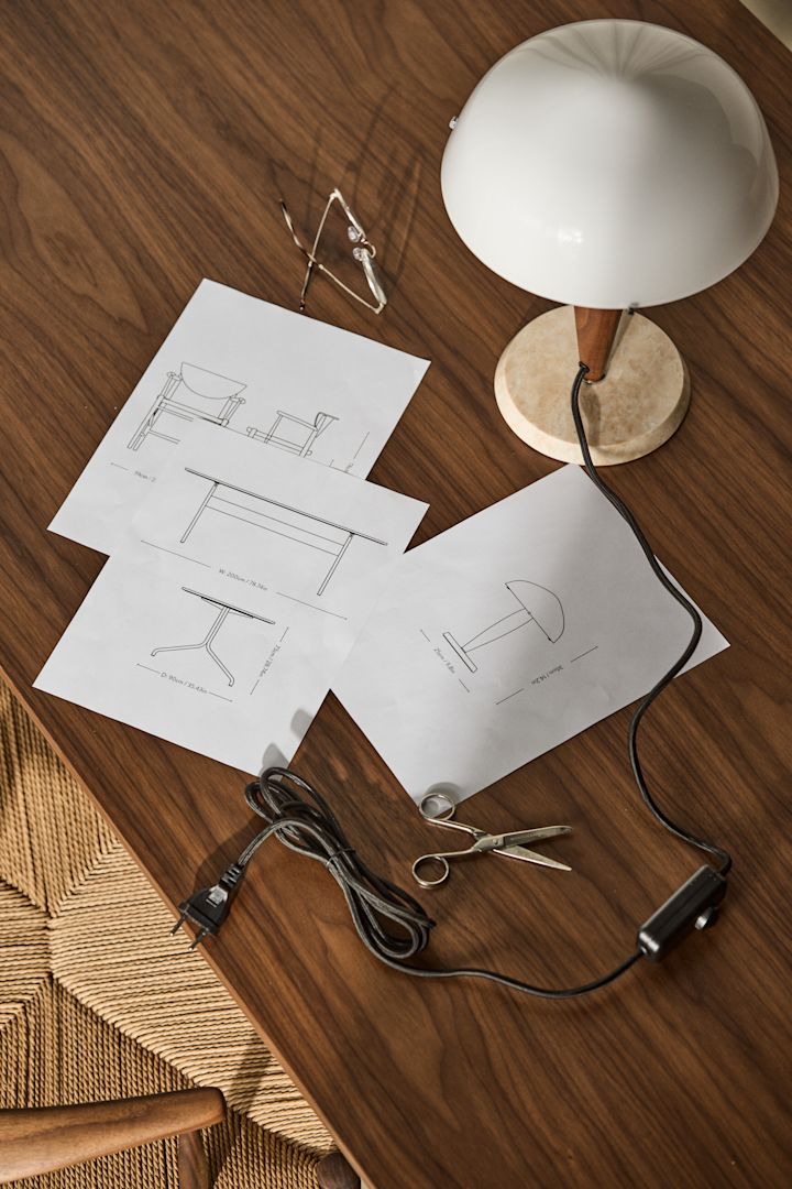 Bild som visar Herman lampa med fot i marmor och valnöt och med skärm i opalvitt glas, här placerad på skrivbord i valnöt med produktskisser utspridda på bordsskivan.