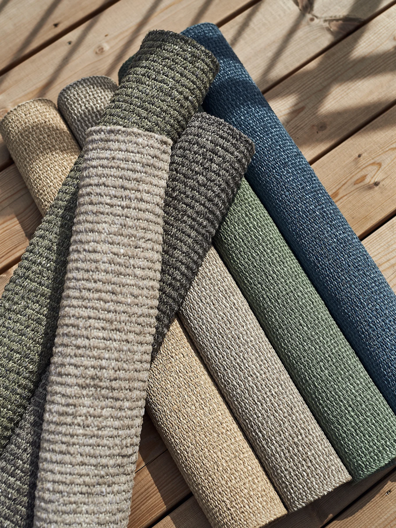 Rullade texturerade mattor i olika naturfärger och jordfärger, inklusive beige, grönt, brunt och blått, ligger på ett trägolv.
