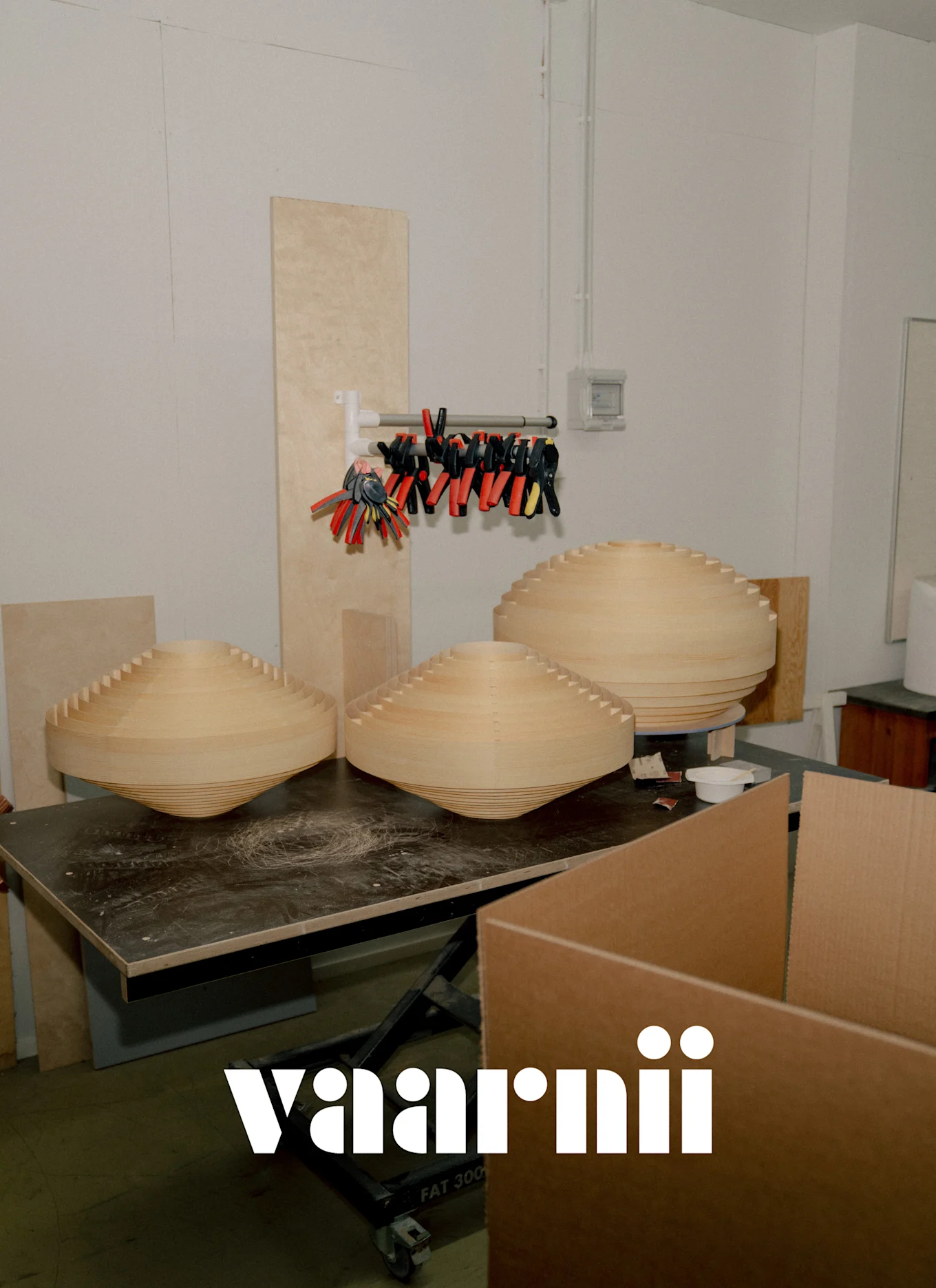 Tre stora, ljusa, lagerdelade träobjekt med konisk form på en mörk arbetsbänk i en Vaarnii-verkstad.