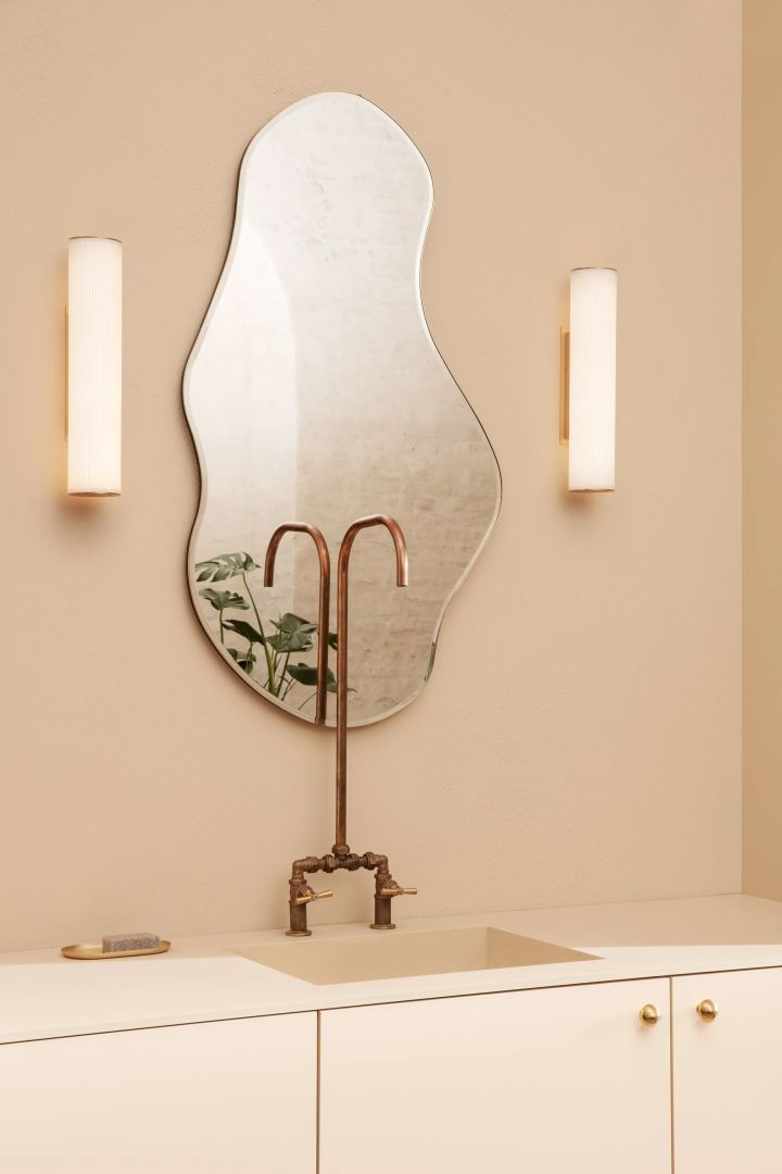 Ett lyxigt badrum kräver en vacker spegel, som på denna bild som visar den organiskt formade spegeln Pond från Ferm Living, placerad på beige badrumsvägg ovanför en vit kommod, på varsin sida av spegeln sitter två tubformade lampor.