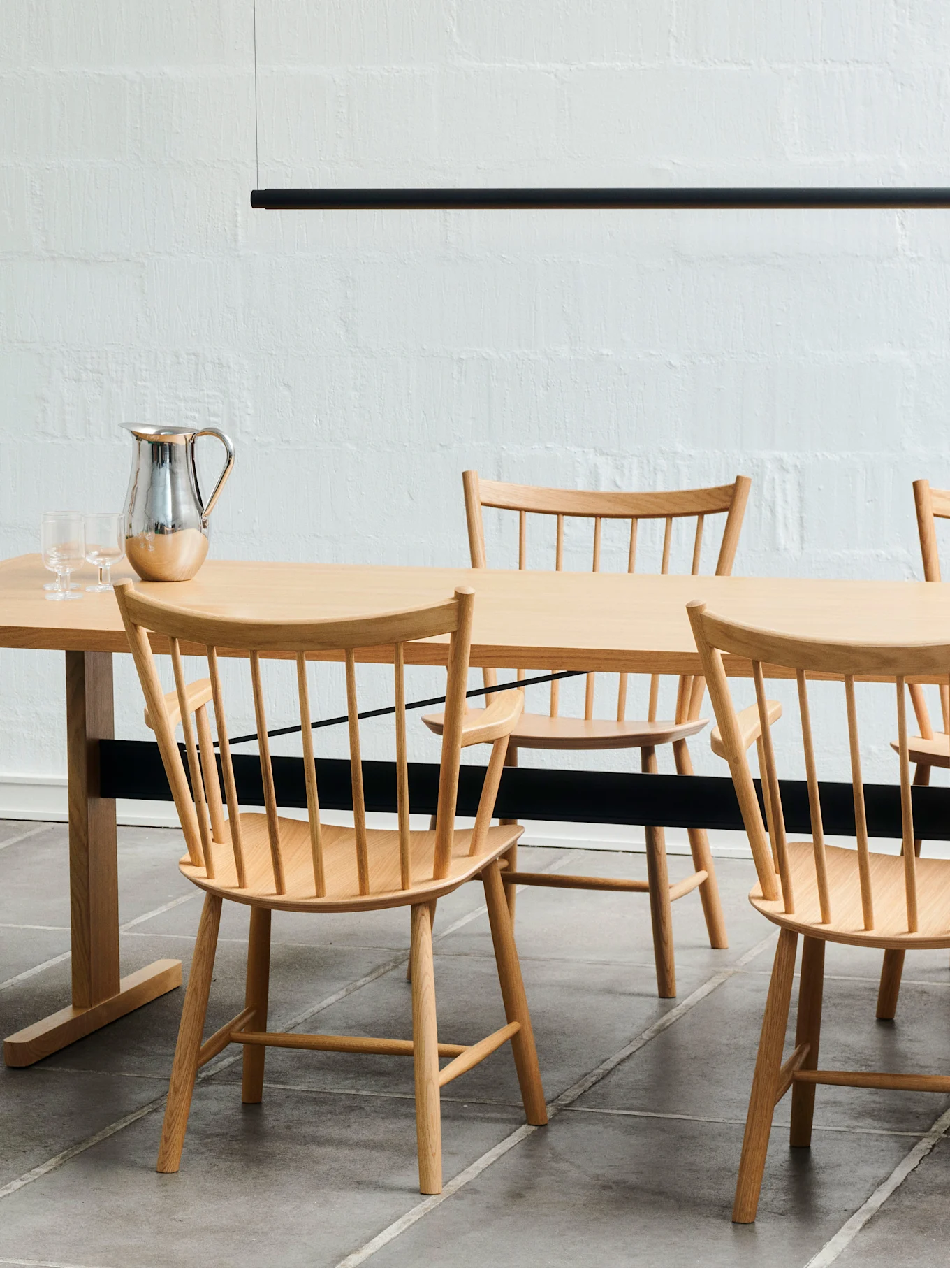 Ett ljust trämatbord med matchande stolar med pinnrygg, en silverkanna och två glas. En svart linjär lampa hänger ovanför.