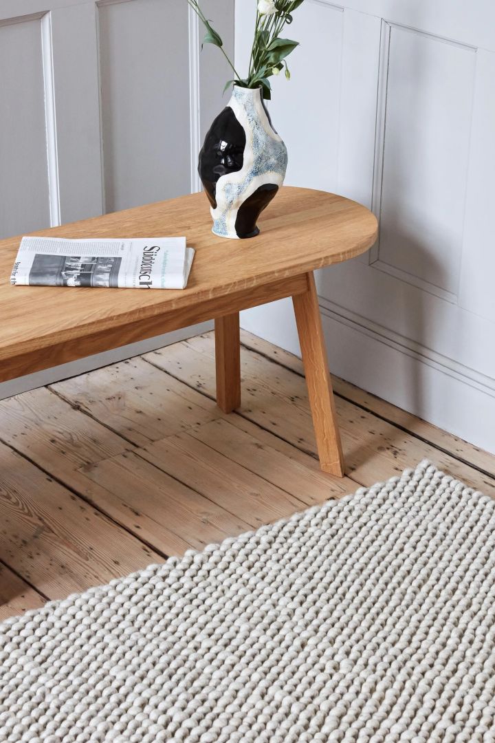 Välj rätt matta, bild som visar Peas ullmatta från HAY i färgen soft grey, här på ett hallgolv i trä med en låg träbänk framför där en vas och tidning är placerade.