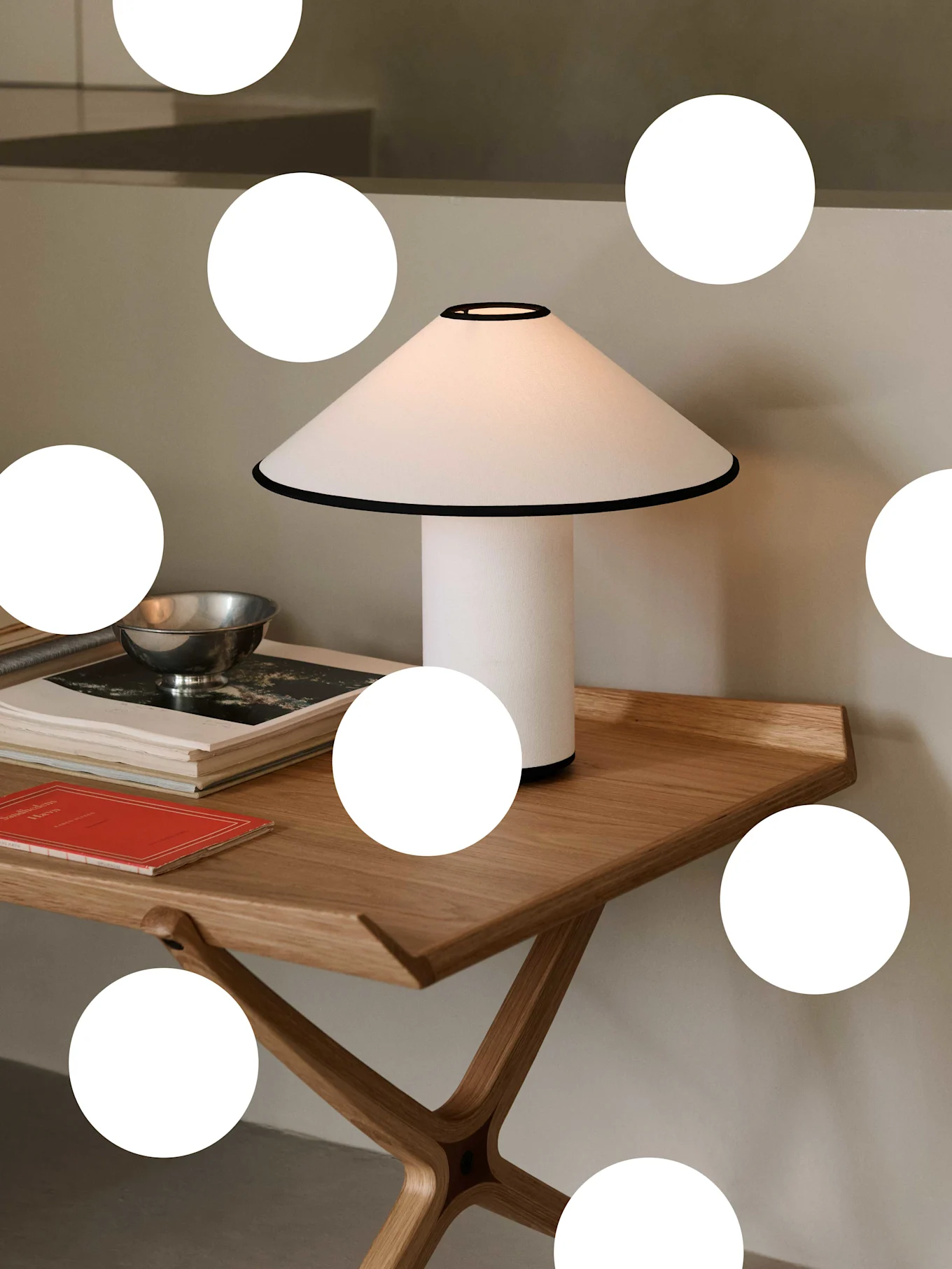 Vit bordslampa med konisk skärm och svart kant på ett ljust träbord, med böcker och en silverskål.