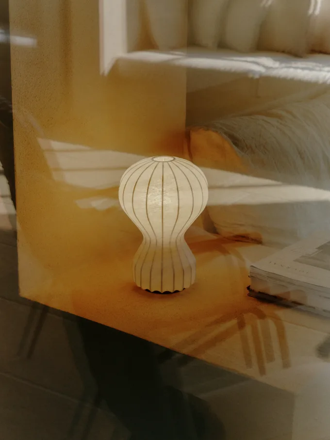 En ljus, räfflad, timglasformad bordslampa på en träyta med ett sovrum i bakgrunden.