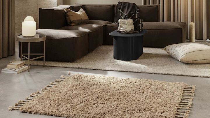 Välj rätt matta, kanske denna härliga ryamatta Amass från Ferm Living, en beige ryamatta med fransar placerad i vardagsrum med brun soffa, betonggolv och stort fönster.