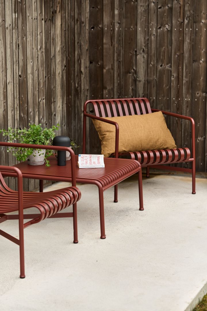 Bild som föreställer Pallisade loungefåtölj och bord i färgen iron red från HAY. Utemöblerna i lackerat stål står placerade mot husvägg på uteplats av betong.