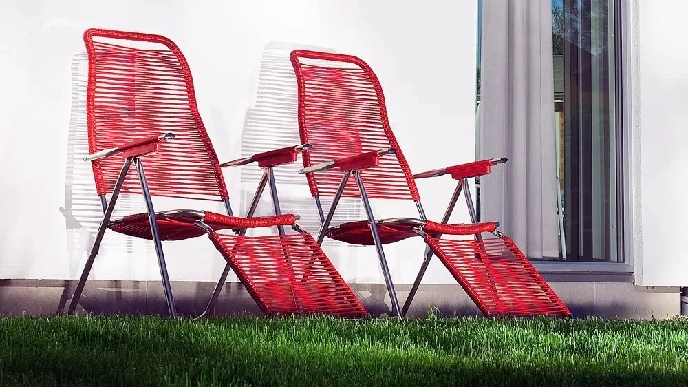 Två Spaghetti solstolar med fotstöd i rött från Fiam står på en gräsmatta mot en vit husvägg