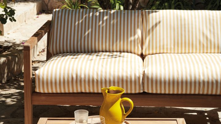 Bild som visar Virkelyst 2-sits loungesoffa från Skagerak. Soffan är tillverkad i ask med mjuka dynor i ett randigt tyg i gult och vitt. framför soffan står ett litet bord i ask med en gul karaff och glas.