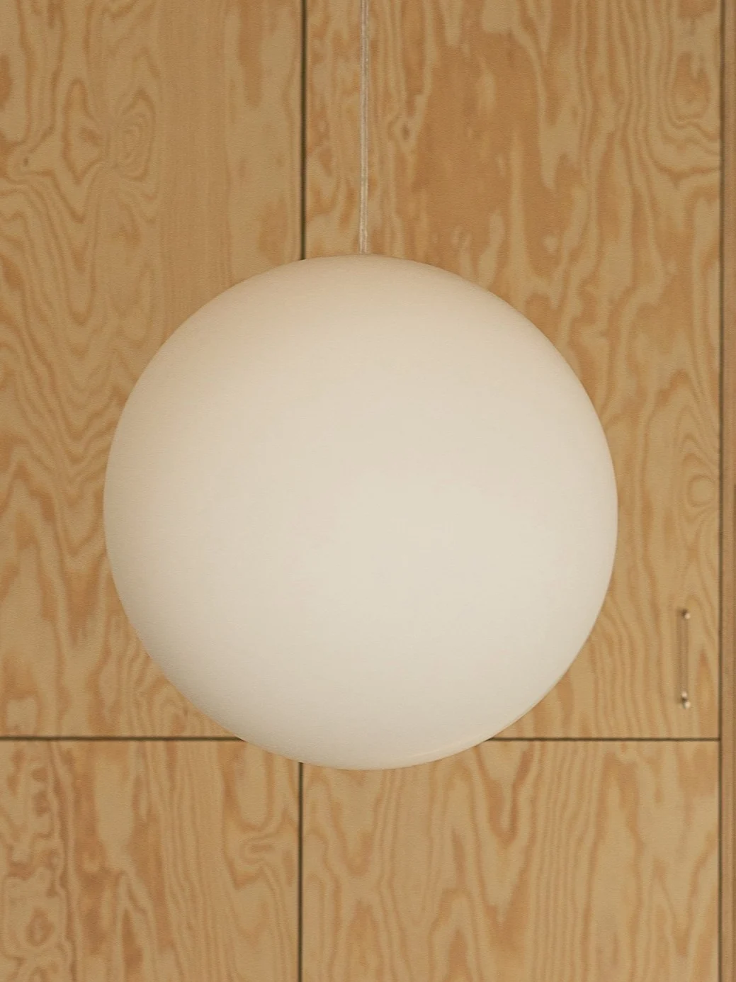 En vit, sfärisk taklampa hänger framför en ljus trävägg med tydlig ådring.