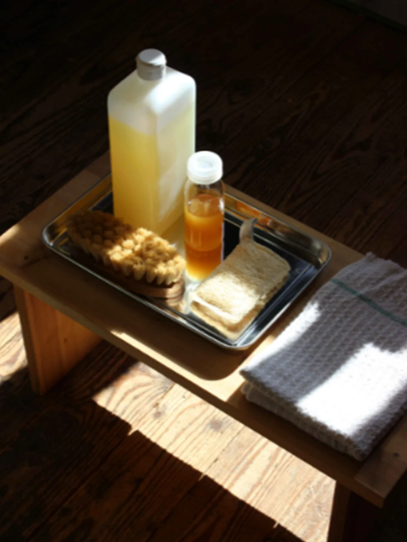 Badartiklar på en bricka: Flaska gul vätska, flaska orange vätska, badborste, loofah. Vit handduk på träbord.