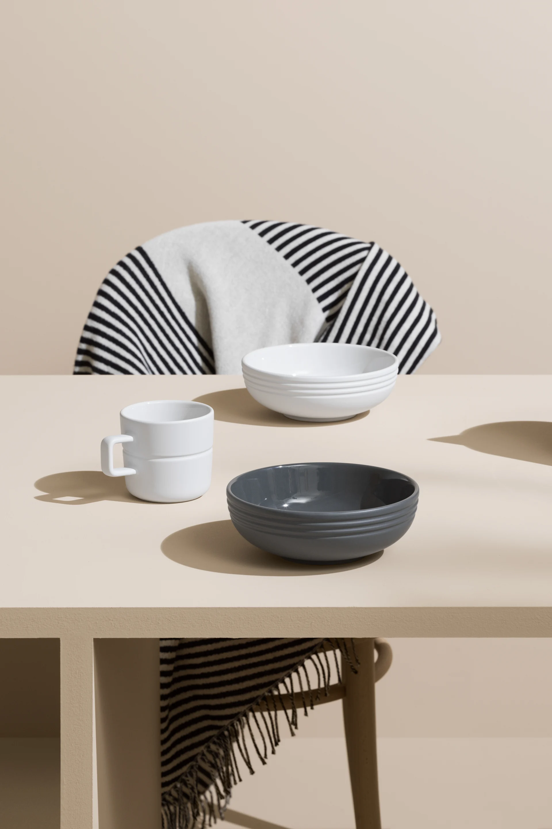 NJRD-porslin skålar i grått och vitt, kaffekopp och en svartvi bomullspläd på stolen. 