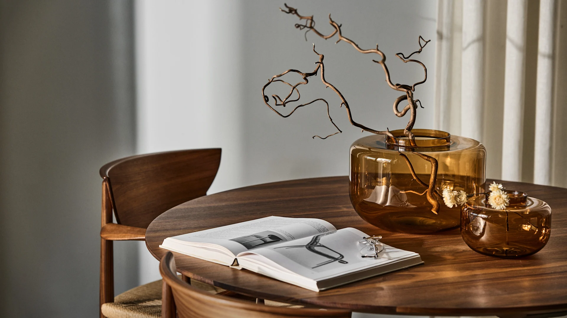 Bild som visar två Drummel-vaser i två storlekar i bärnstensgult placerade på mörkt träbord med mörka trästolar. I den största vasen är grenar av trollhassel placerade.