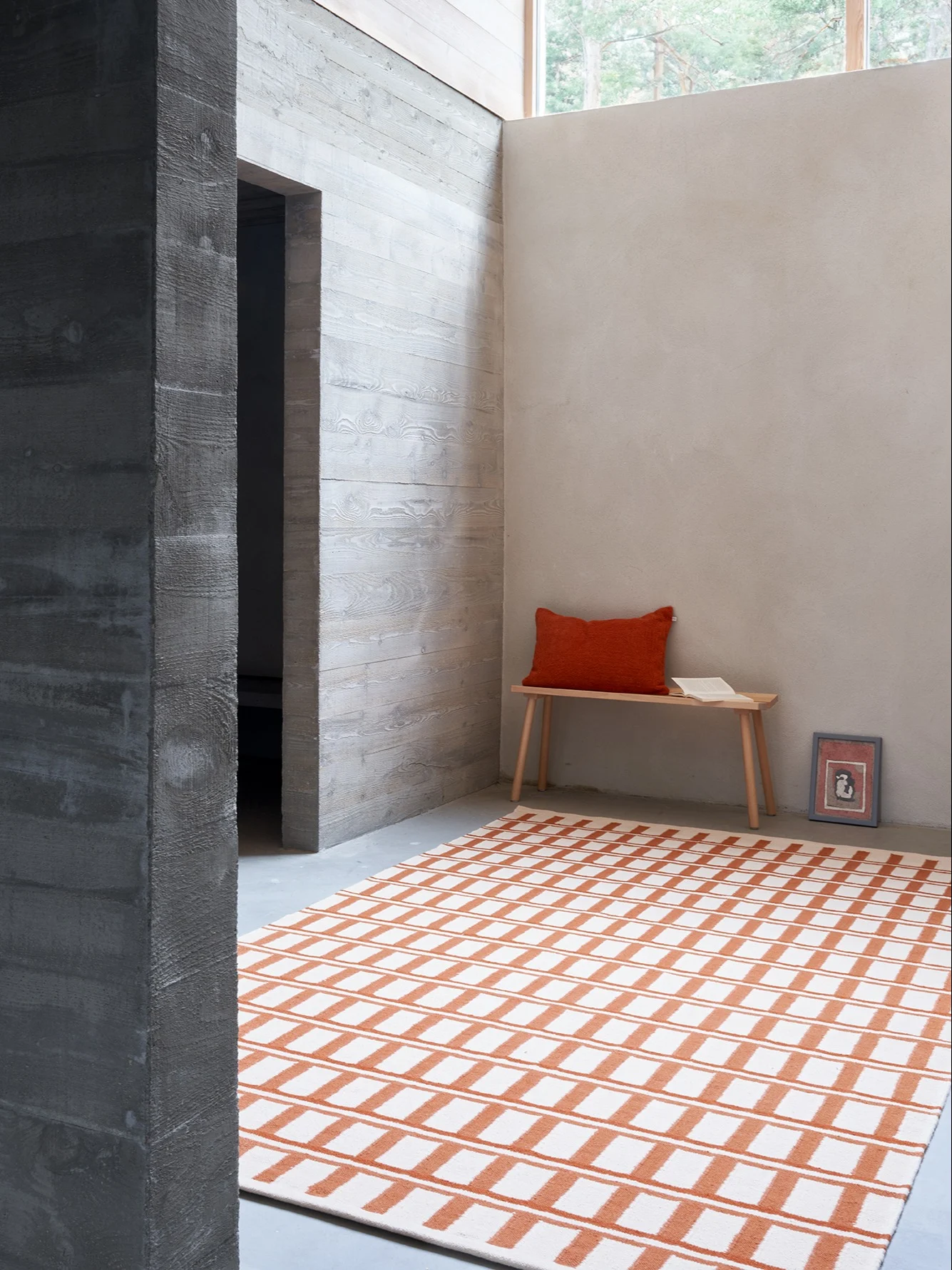 Ett rum med en vit- och orangerandig matta, en träbänk med orange kudde, en bok och en bild. Betongväggar.