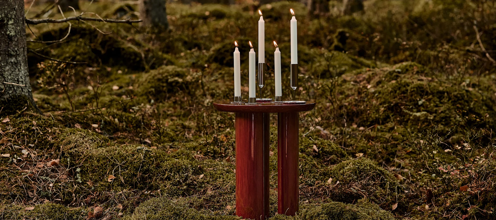 Julinspiration till vardagsrummet, här med Tung JA3 sidobord i rött från &tradition med en grupp levande ljus på, placerat i en skog klädd av grön mossa.