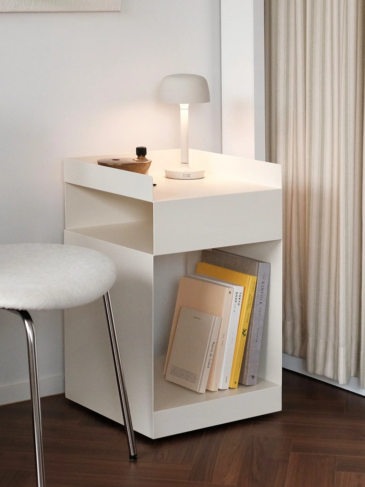 Modernt hörn med vit vägg, ljust abstrakt måleri, stol, sidobord med lampa, grön hängväxt och randiga gardiner.