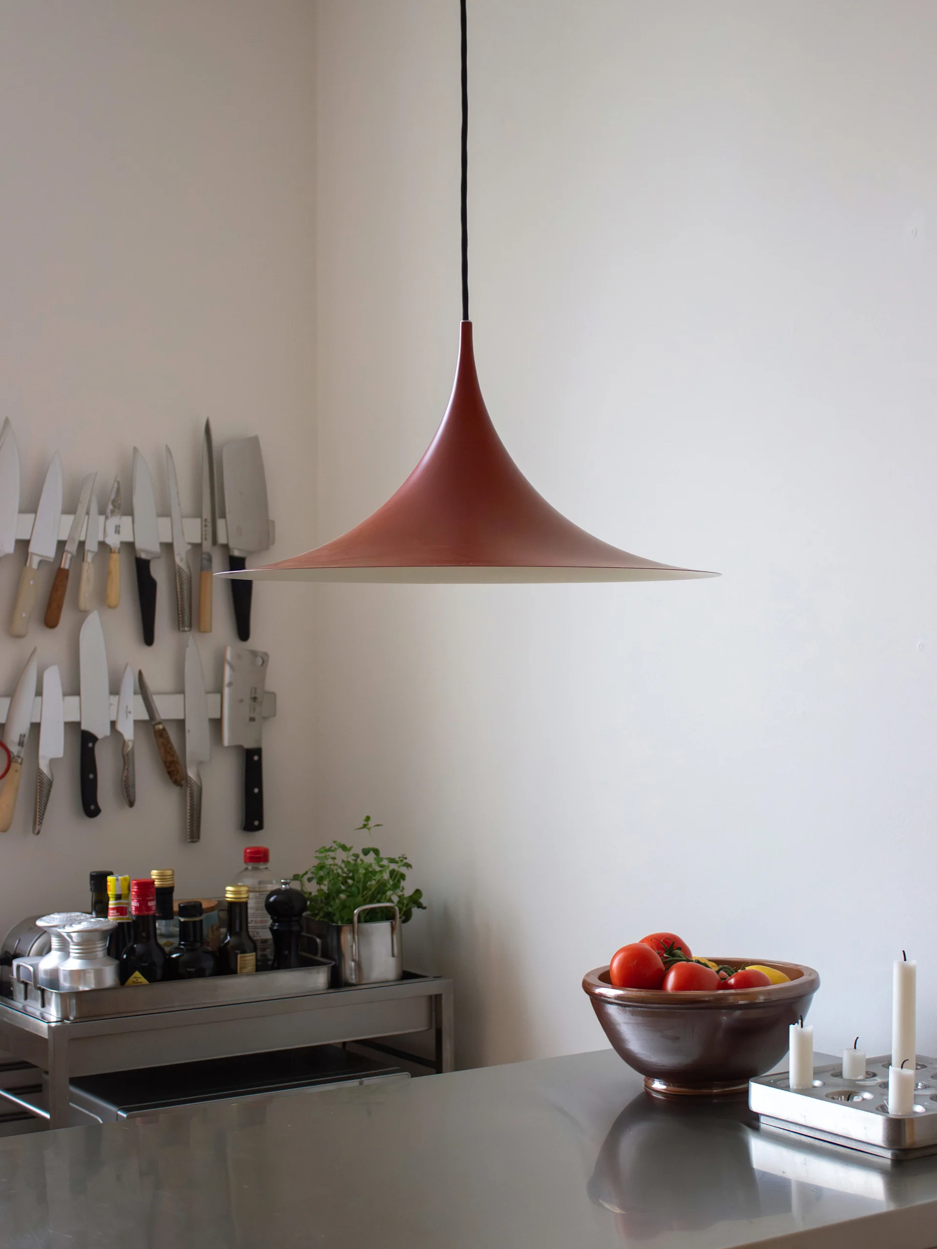 Modernt kök: rödbrun klockformad lampa, bänkskiva i rostfritt stål med tomater och ljus, vägg med hängande knivar.