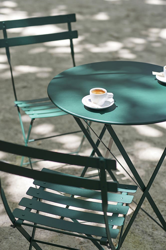 Miljöbild föreställandes bistro cafébord och stolar i färgen cedar green. ståendes på en grusplätt och har två kaffekoppar på sig. 