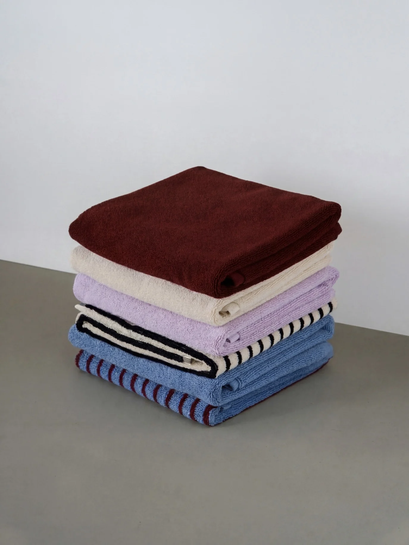 En stapel med fem vikta handdukar i olika färger: vinrött, gräddvitt, lavendel, randigt svart/vitt och randigt blått/vinrött.