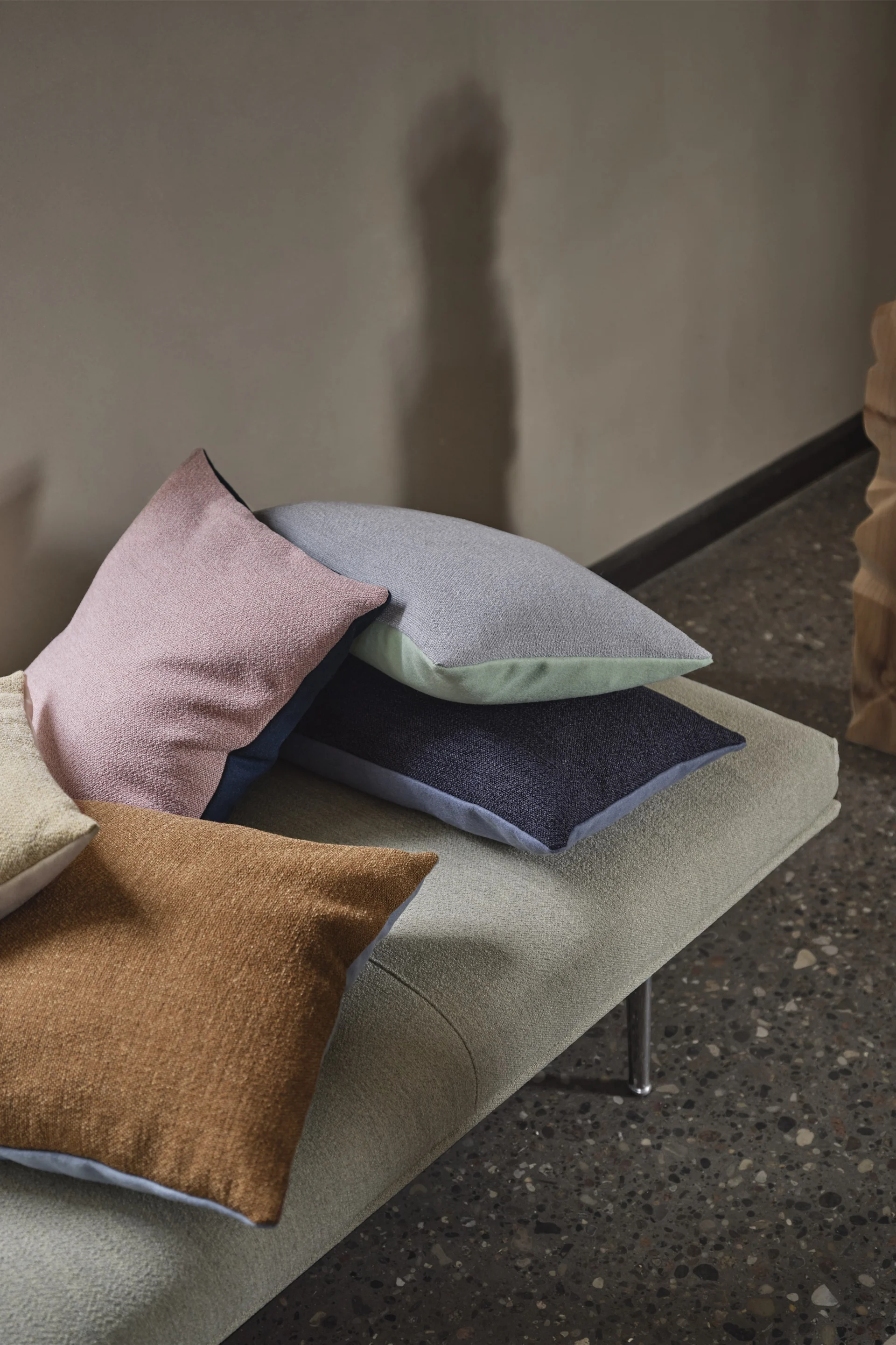 Bild som visar flera Mingle kuddar från Muuto i olika dova färger utspridda på en divan intill ett fönster.