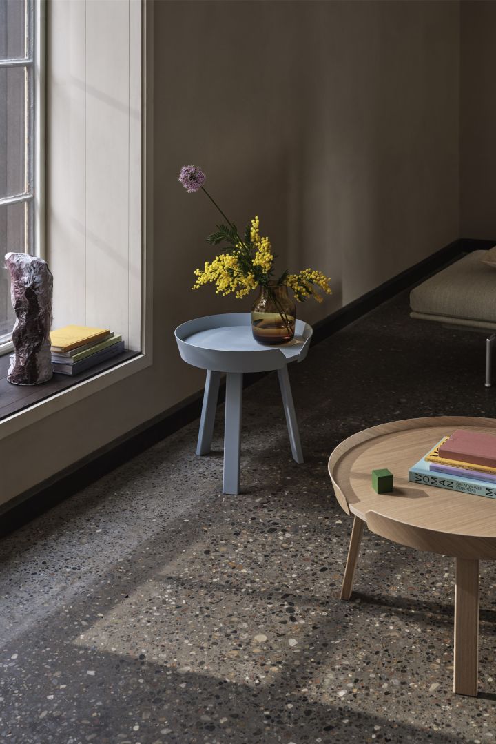 Bild som visar två stycken Around bord från Muuto, två runda soff- och sidobord i ek och ett i ljusblått placerade i ett rum med grått terazzo-golv. På det lilla ljusblå bordet står en gul vas med en gul blombukett.