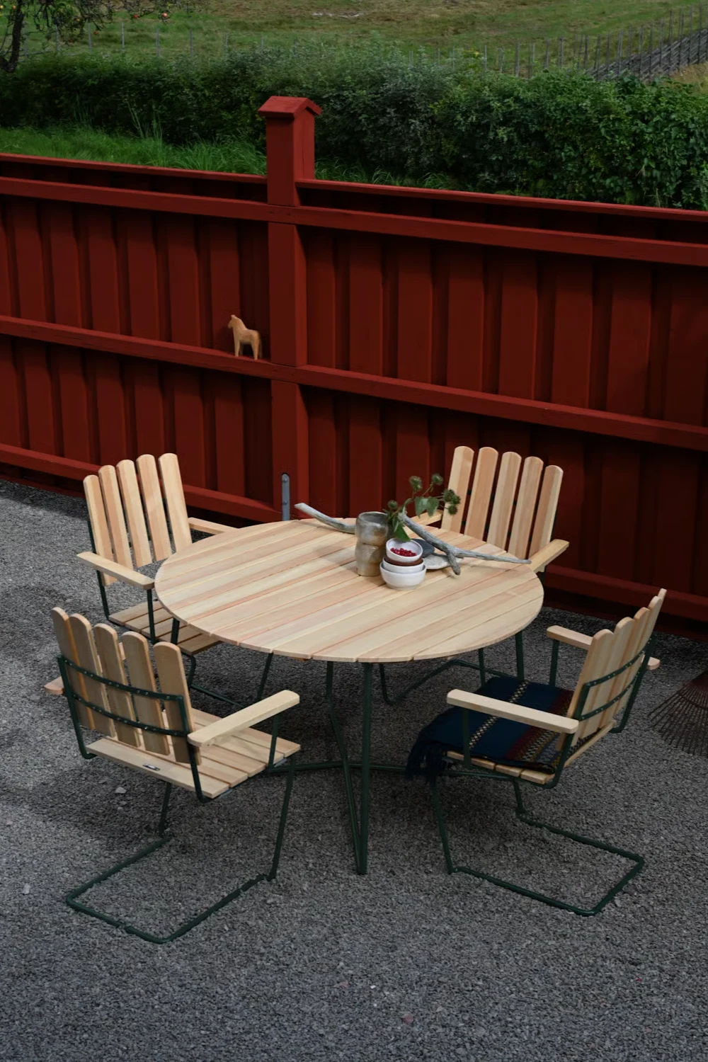 Runt träbord och fyra stolar på en grusbelagd uteplats framför ett rött staket med en trähäst.